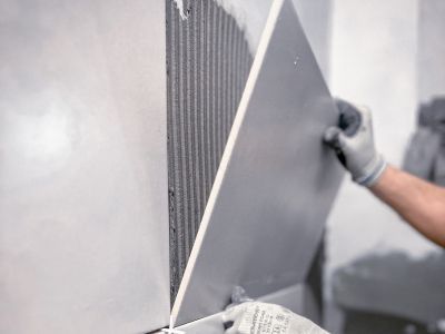 Person setzt große graue Wandfliese mit Kleber ein, trägt Handschuhe, präzise Arbeit bei Fliesenverlegung sichtbar. Person setzt große graue Wandfliese mit Kleber ein, trägt Handschuhe, präzise Arbeit bei Fliesenverlegung sichtbar.